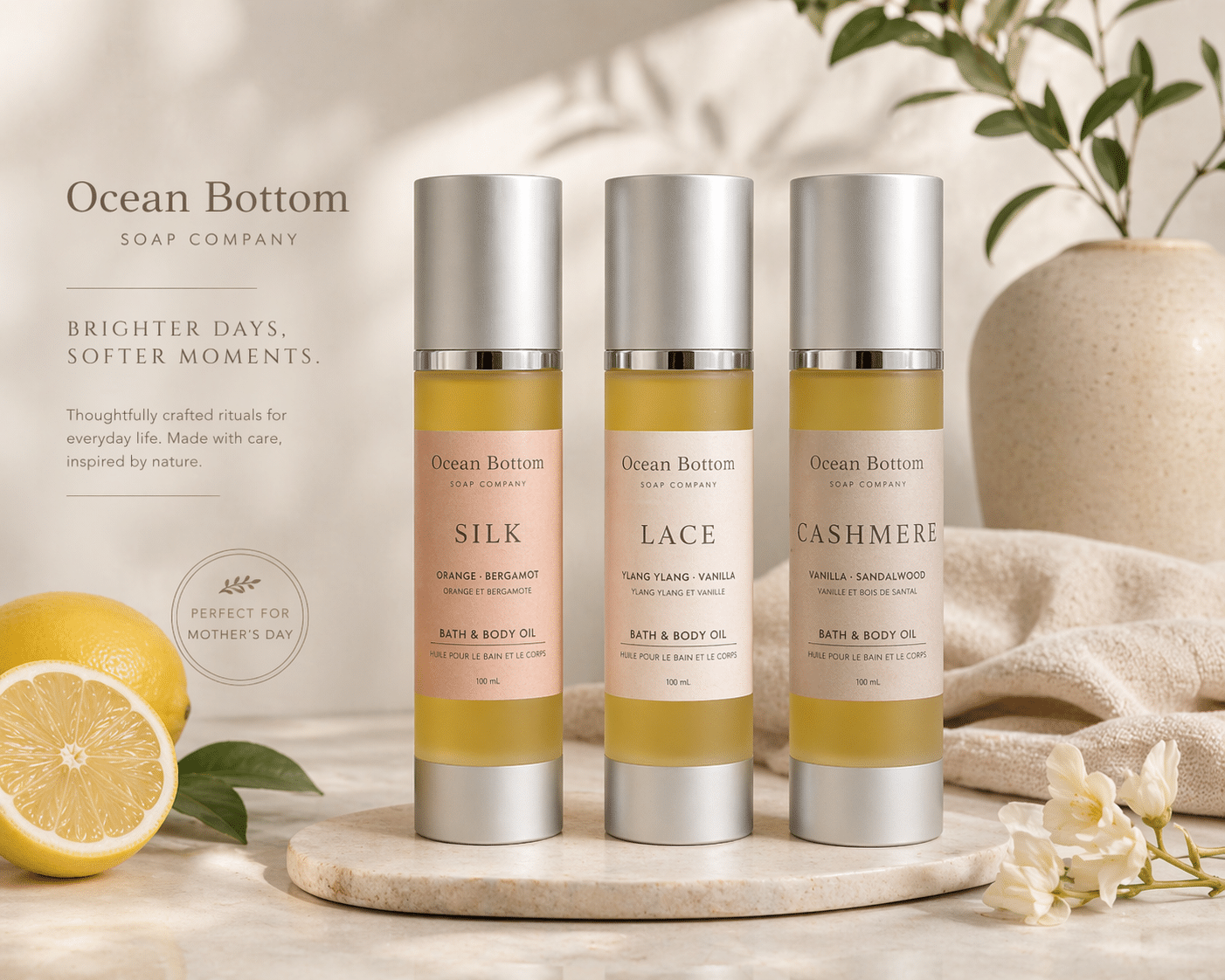Three Ocean Bottom bath & body oil bottles (Silk, Lace, Cashmere) on a round stone platform with a lemon and green plants in a soft beige background.